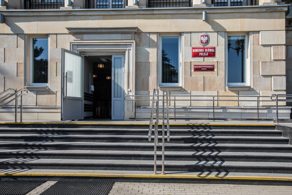 Wejście główne do Komendy Głównej Policji. Widoczne schody, drzwi. Po prawej stronie drzwi, na budynku umieszczona jest czerwona tablica z napisem „Komenda Główna Policji” oraz godło państwowe.