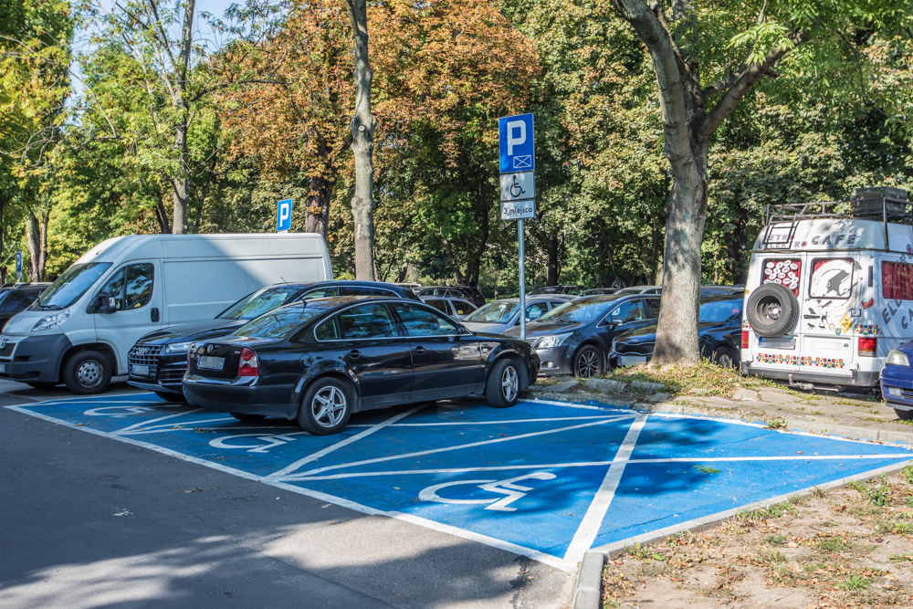 Trzy miejsca parkingowe przeznaczone dla osób z niepełnosprawnościami oznaczone niebieską kopertą i pionowym znakiem drogowym. 
